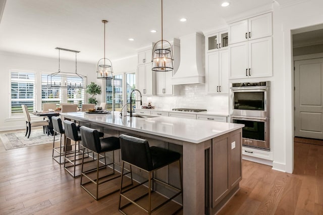 kitchen with sink, premium range hood, white cabinetry, stainless steel appliances, and a center island with sink