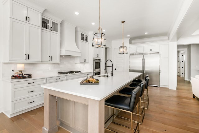 kitchen with appliances with stainless steel finishes, decorative light fixtures, white cabinets, a large island with sink, and custom range hood