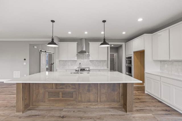 kitchen with a spacious island, wall chimney range hood, pendant lighting, a barn door, and white cabinetry