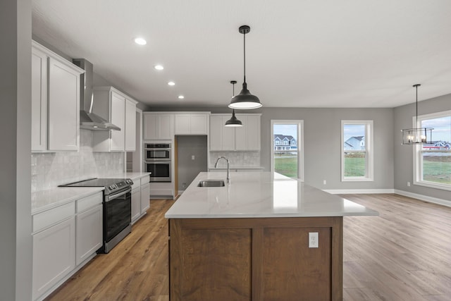 kitchen with stainless steel appliances, sink, wall chimney range hood, white cabinets, and an island with sink