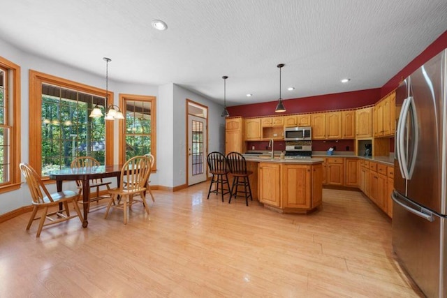 kitchen featuring a kitchen island with sink, light wood-style floors, light countertops, appliances with stainless steel finishes, and decorative light fixtures