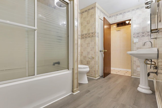 bathroom with wood-type flooring, tile walls, shower / bath combination with glass door, and toilet