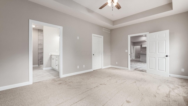 unfurnished bedroom with a raised ceiling, light colored carpet, ensuite bath, and ceiling fan