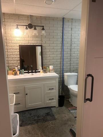 bathroom featuring a paneled ceiling, vanity, brick wall, and toilet