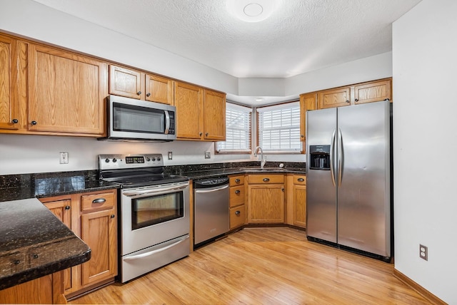 kitchen with light wood-type flooring, a textured ceiling, stainless steel appliances, sink, and dark stone countertops