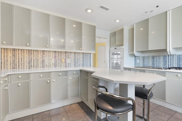 kitchen with kitchen peninsula, light tile patterned floors, a kitchen bar, gray cabinetry, and double oven