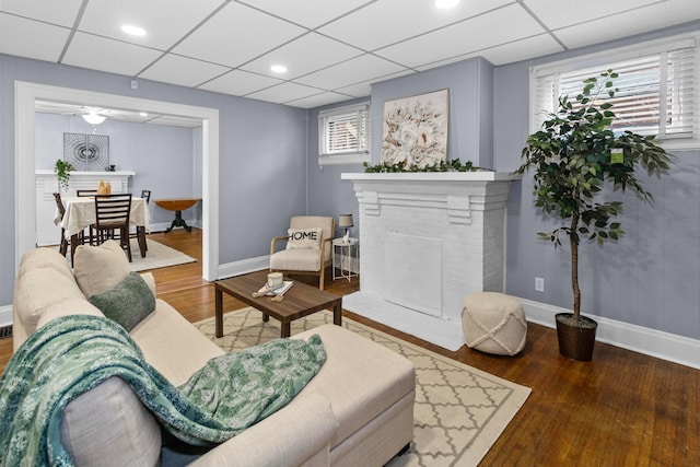 living area featuring a brick fireplace, a drop ceiling, baseboards, and wood finished floors