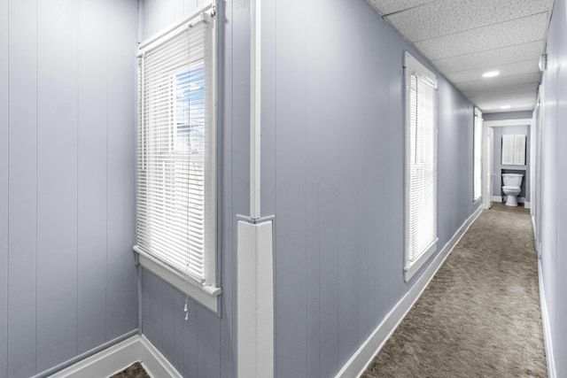 hallway featuring baseboards, wooden walls, dark carpet, and a drop ceiling