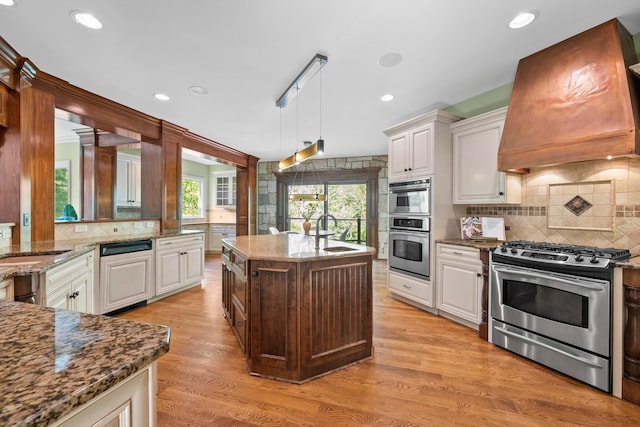 kitchen with appliances with stainless steel finishes, custom exhaust hood, decorative light fixtures, stone counters, and light hardwood / wood-style floors