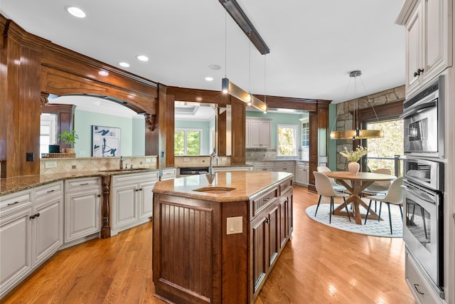 kitchen featuring stainless steel oven, tasteful backsplash, pendant lighting, a kitchen island with sink, and light wood-type flooring