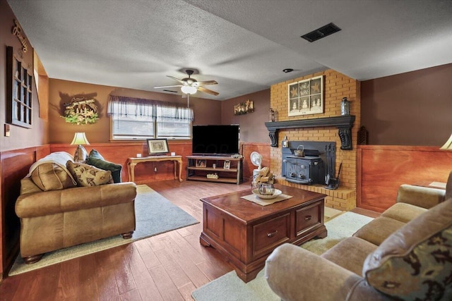 living area with wainscoting, ceiling fan, wood finished floors, a wood stove, and a textured ceiling