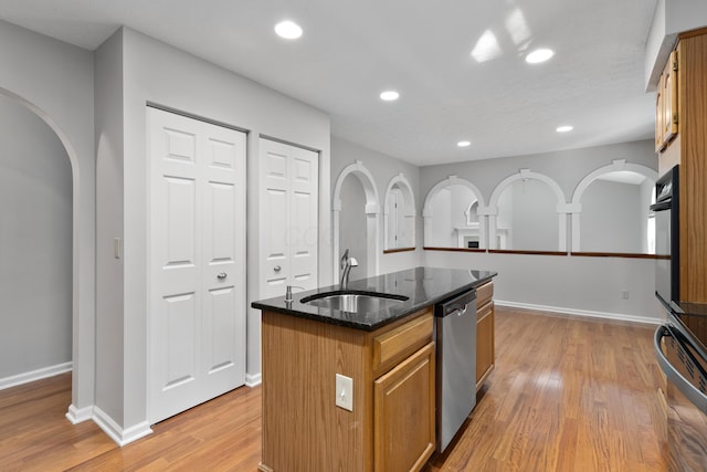 kitchen featuring sink, stainless steel dishwasher, an island with sink, dark stone counters, and light wood-type flooring