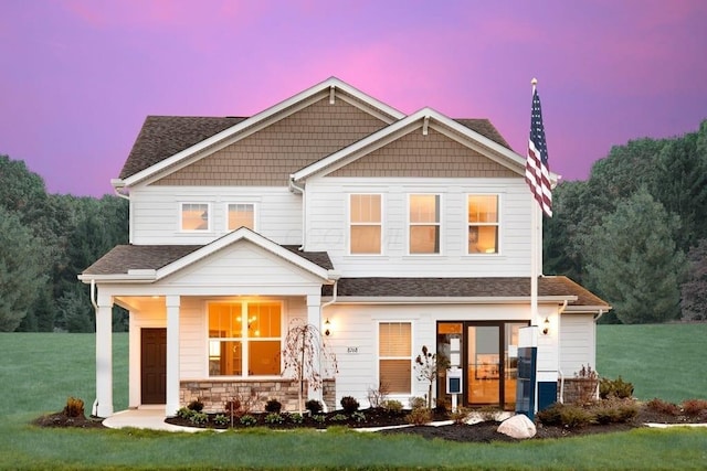 view of front of home featuring a yard, roof with shingles, stone siding, and covered porch