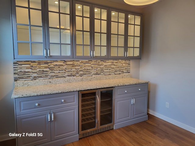 bar featuring dark wood-type flooring, decorative backsplash, gray cabinets, light stone countertops, and beverage cooler