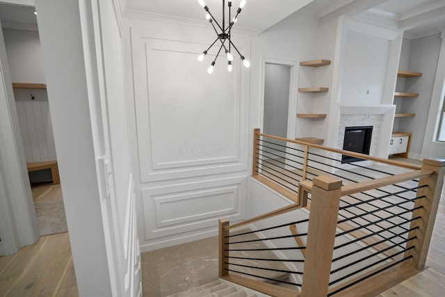 stairway with an inviting chandelier, a stone fireplace, hardwood / wood-style flooring, built in shelves, and ornamental molding