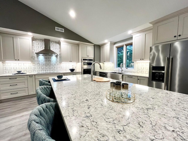 kitchen featuring vaulted ceiling, wall chimney exhaust hood, decorative backsplash, light wood-type flooring, and stainless steel appliances