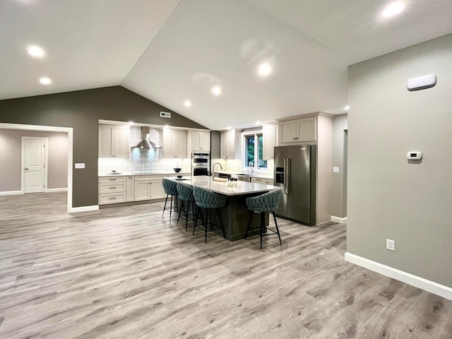 kitchen with appliances with stainless steel finishes, a kitchen breakfast bar, a kitchen island with sink, wall chimney range hood, and light hardwood / wood-style floors