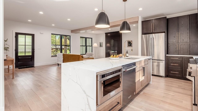 kitchen featuring stainless steel appliances, light stone counters, an island with sink, light hardwood / wood-style floors, and decorative light fixtures