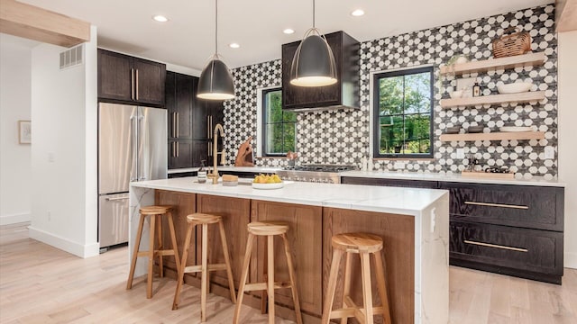 kitchen with appliances with stainless steel finishes, tasteful backsplash, decorative light fixtures, light hardwood / wood-style floors, and an island with sink