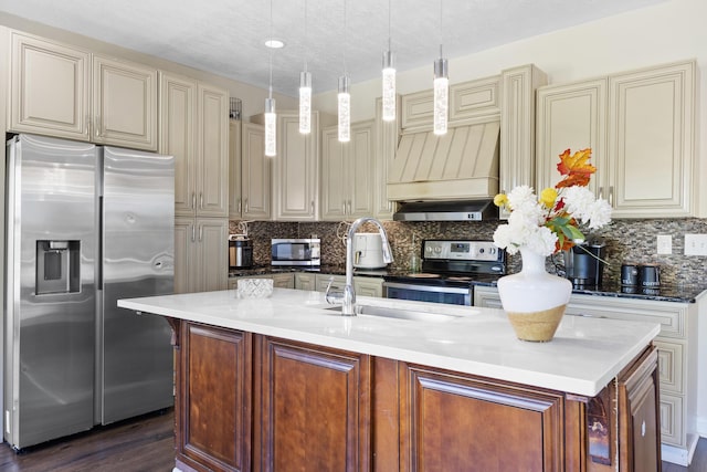 kitchen with dark wood-type flooring, pendant lighting, cream cabinetry, a kitchen island with sink, and appliances with stainless steel finishes