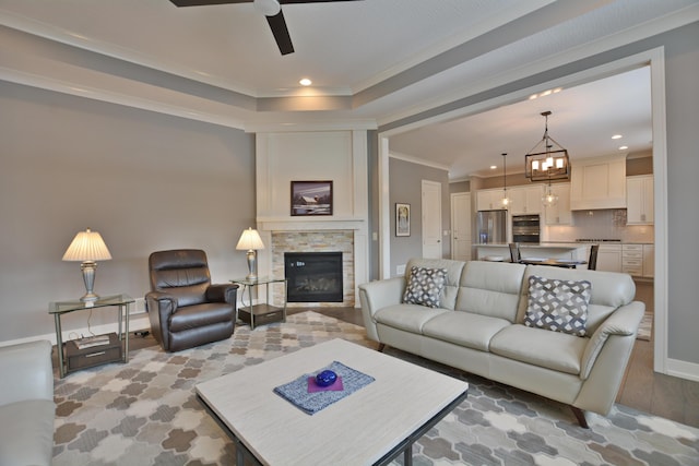 living room with ornamental molding, light wood-type flooring, ceiling fan with notable chandelier, a tray ceiling, and a stone fireplace