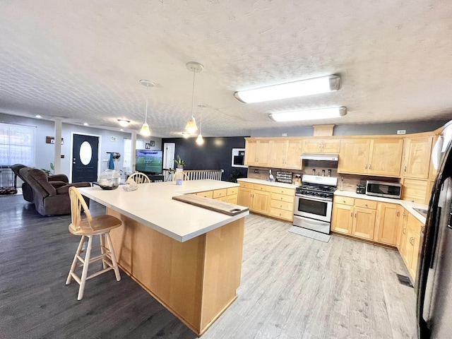 kitchen with light brown cabinetry, a textured ceiling, stainless steel appliances, decorative light fixtures, and light hardwood / wood-style flooring