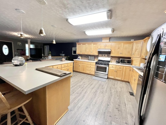 kitchen with light brown cabinets, stainless steel appliances, pendant lighting, a textured ceiling, and light hardwood / wood-style floors