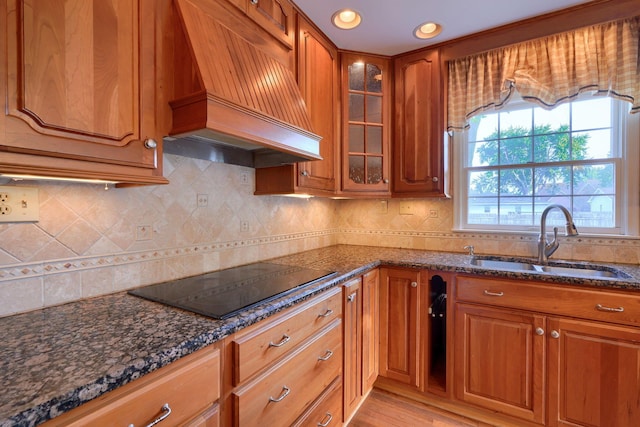 kitchen with black electric stovetop, custom range hood, sink, light hardwood / wood-style flooring, and dark stone countertops