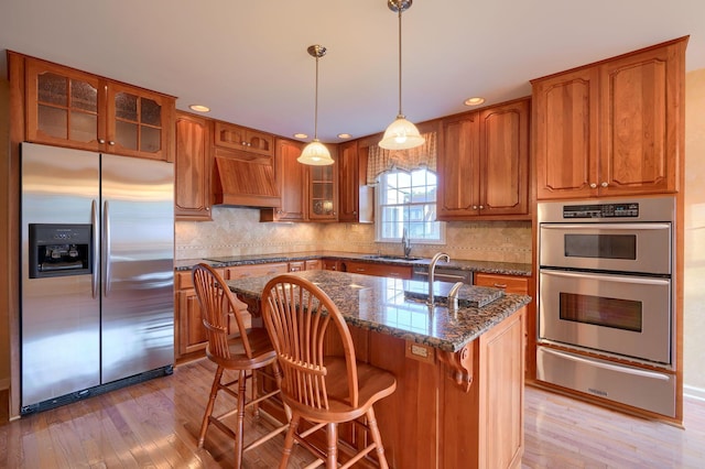 kitchen with custom range hood, stainless steel appliances, a center island with sink, light hardwood / wood-style flooring, and dark stone countertops