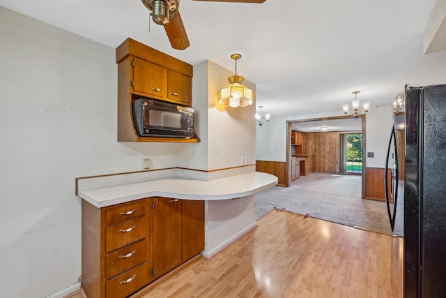 kitchen with hanging light fixtures, light hardwood / wood-style flooring, wooden walls, black appliances, and ceiling fan with notable chandelier