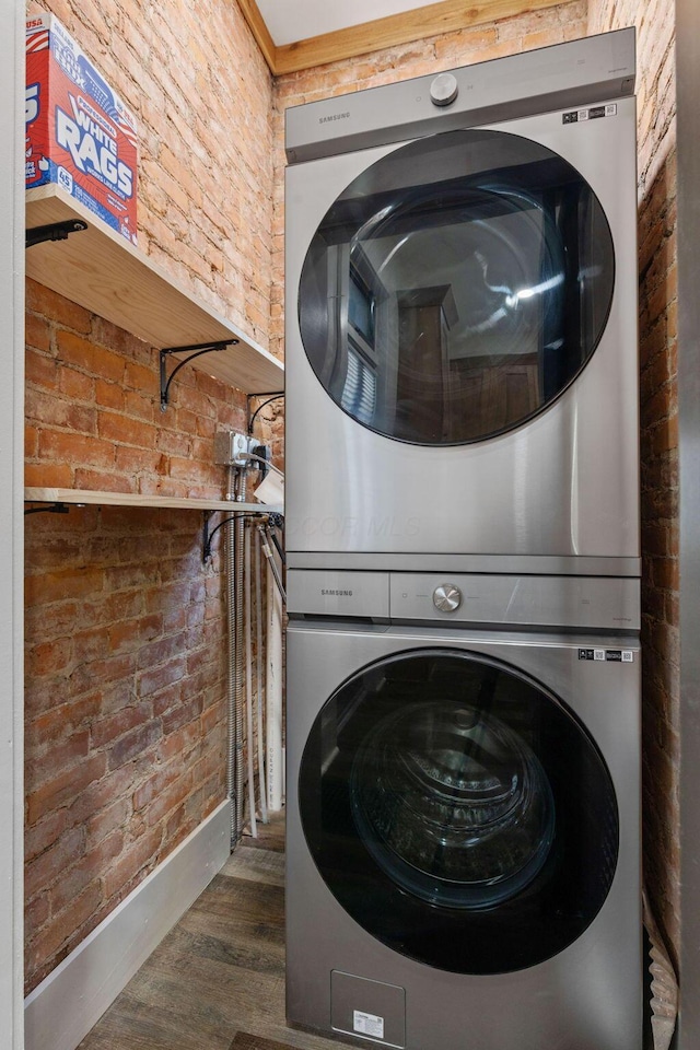 clothes washing area with stacked washer / dryer, dark hardwood / wood-style flooring, and brick wall