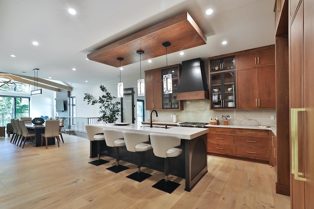 kitchen with premium range hood, sink, light wood-type flooring, an island with sink, and decorative light fixtures