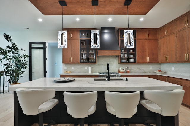 kitchen featuring light hardwood / wood-style floors, custom range hood, a kitchen island with sink, and tasteful backsplash