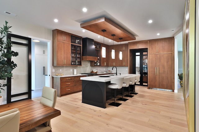 kitchen with a center island with sink, custom exhaust hood, hanging light fixtures, and light hardwood / wood-style flooring