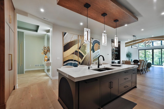 kitchen with sink, light hardwood / wood-style flooring, an island with sink, decorative light fixtures, and light stone counters