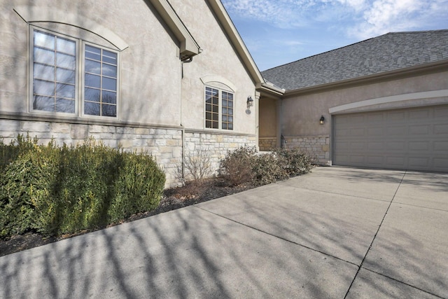 view of side of property featuring driveway, a garage, stone siding, roof with shingles, and stucco siding