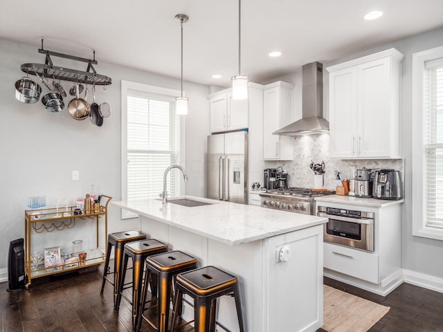 kitchen with a healthy amount of sunlight, wall chimney exhaust hood, stainless steel appliances, and an island with sink