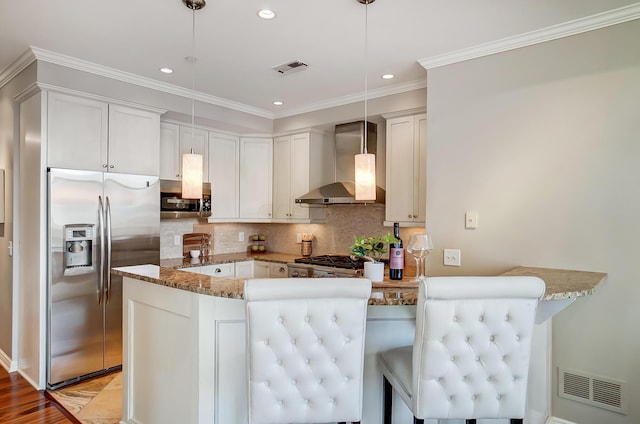 kitchen with wall chimney range hood, kitchen peninsula, appliances with stainless steel finishes, decorative light fixtures, and white cabinetry