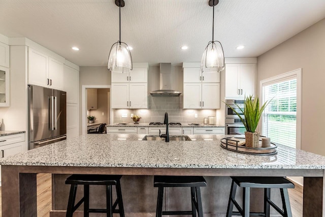 kitchen with appliances with stainless steel finishes, a kitchen island with sink, wall chimney range hood, light hardwood / wood-style flooring, and white cabinets