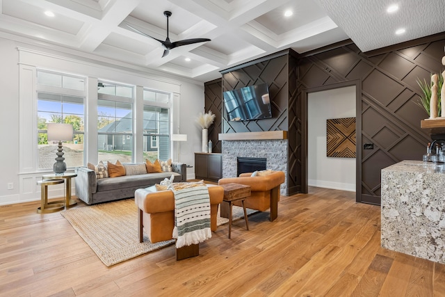 living room with beamed ceiling, light hardwood / wood-style floors, a stone fireplace, and ceiling fan