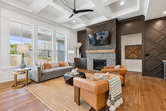 living room with coffered ceiling, a stone fireplace, light hardwood / wood-style flooring, ceiling fan, and beamed ceiling