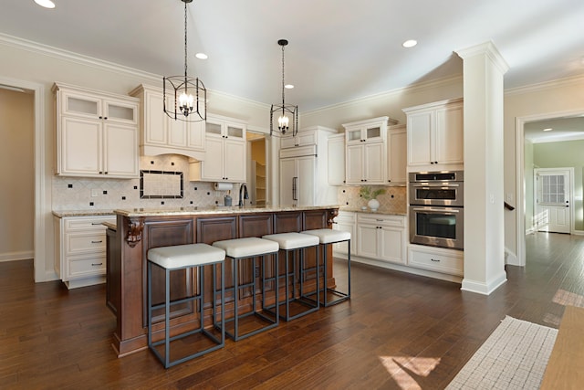 kitchen with light stone counters, hanging light fixtures, paneled built in refrigerator, double oven, and an island with sink