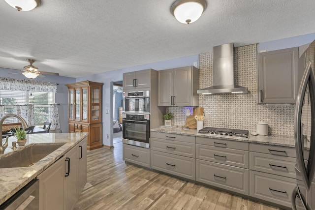 kitchen featuring wall chimney range hood, sink, light stone countertops, light wood-type flooring, and stainless steel appliances