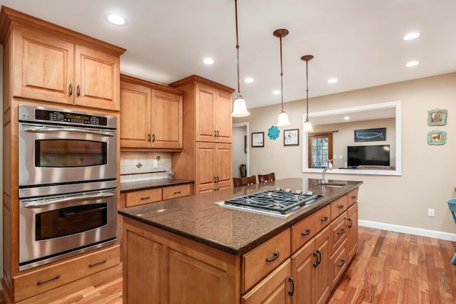 kitchen featuring appliances with stainless steel finishes, pendant lighting, an island with sink, backsplash, and light hardwood / wood-style flooring