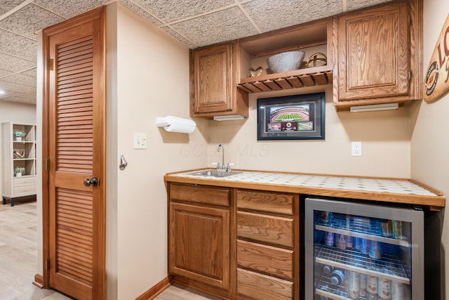 bar featuring sink, light hardwood / wood-style floors, a drop ceiling, tile countertops, and beverage cooler