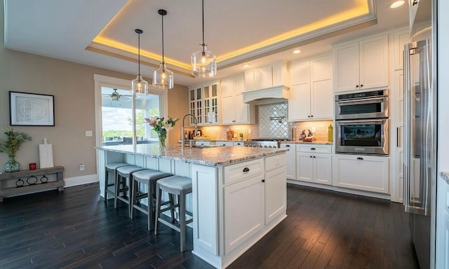 kitchen with a raised ceiling, a center island with sink, white cabinets, and dark wood-type flooring