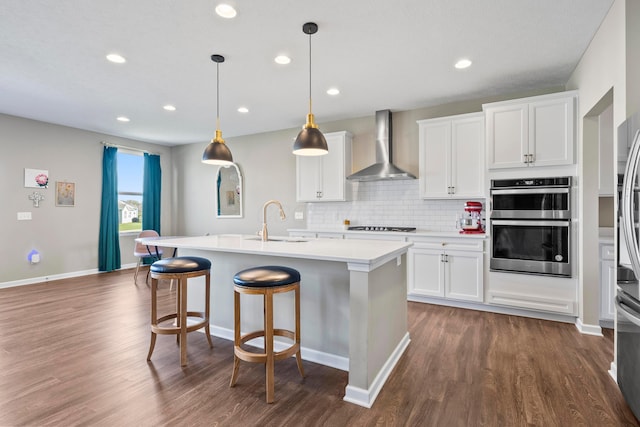 kitchen featuring white cabinets, wall chimney exhaust hood, an island with sink, and decorative light fixtures