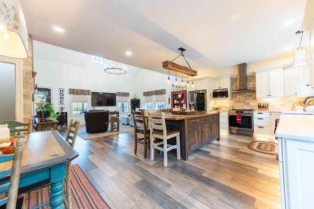 kitchen featuring appliances with stainless steel finishes, wall chimney range hood, high vaulted ceiling, light hardwood / wood-style floors, and white cabinetry