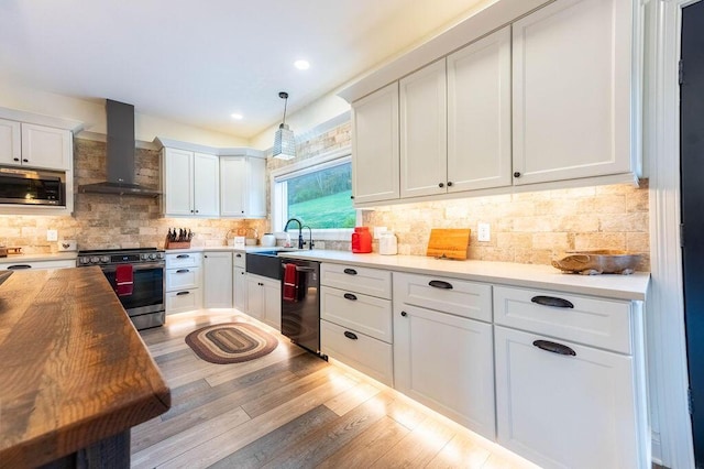 kitchen with pendant lighting, butcher block counters, wall chimney exhaust hood, appliances with stainless steel finishes, and white cabinetry