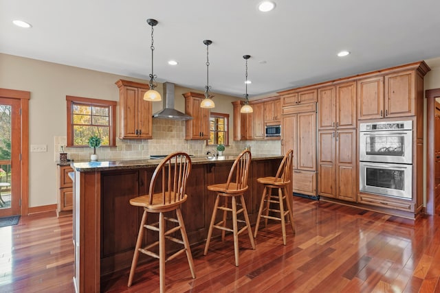 kitchen featuring wall chimney exhaust hood, built in appliances, dark stone countertops, decorative light fixtures, and a kitchen island